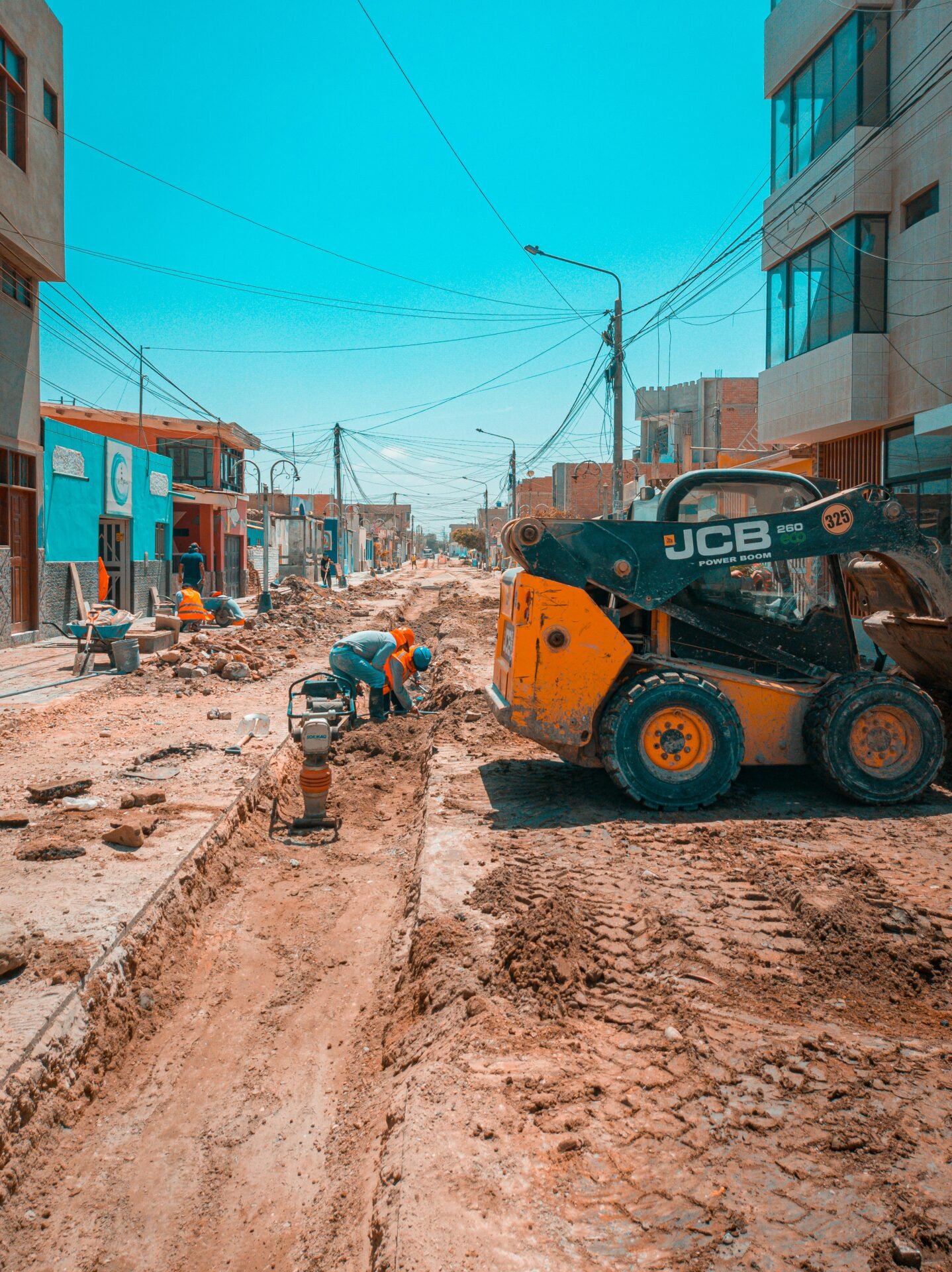 Busy urban road construction scene with workers and construction machinery under a bright blue sky.